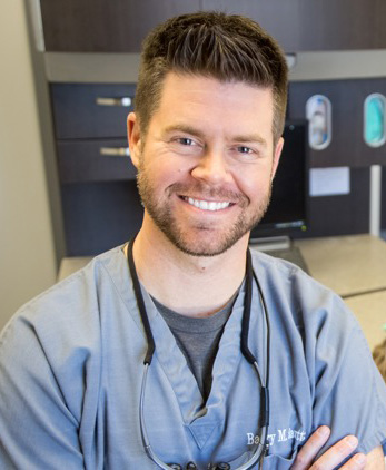 The image is a photograph of a man in a dental office, smiling at the camera. He has short hair, facial stubble, and appears to be wearing scrubs, suggesting he may work in healthcare or dentistry.