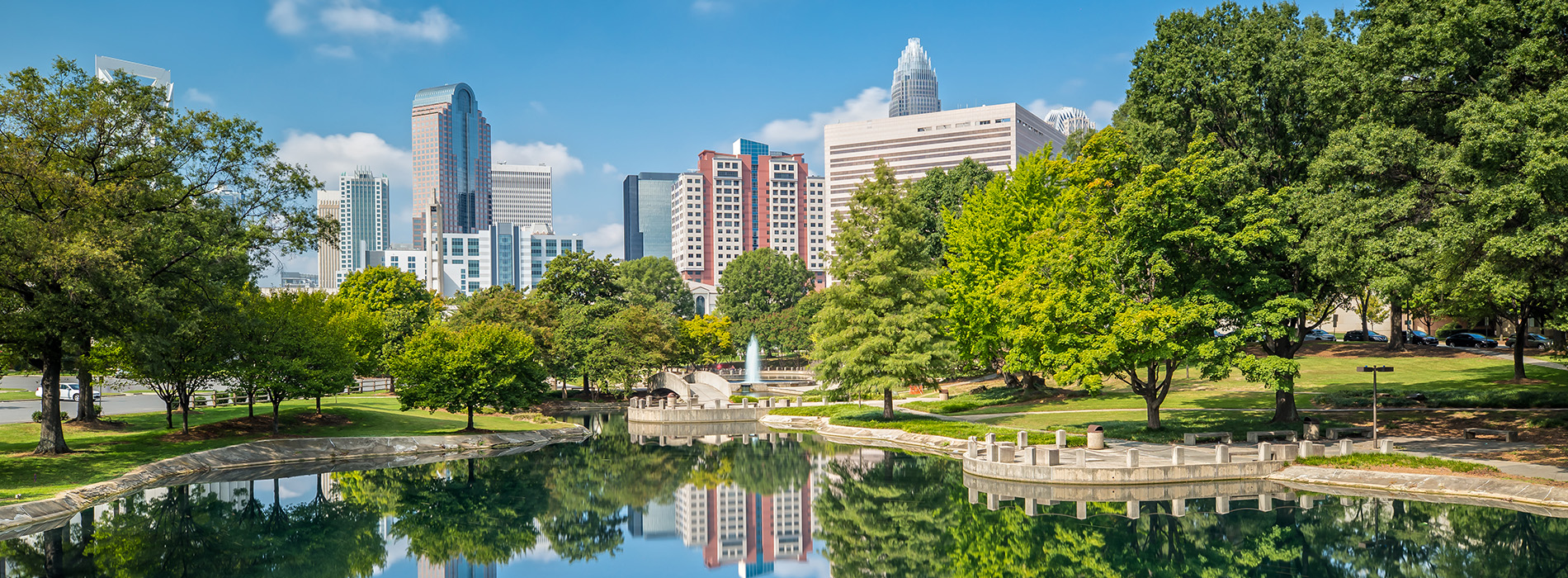 The image is a photograph of an urban park scene on a sunny day, featuring a large pond with a reflection of trees and a city skyline in the background.