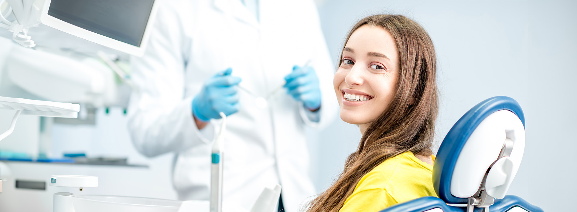 A woman in a dental chair is smiling at the camera, with a dentist standing behind her, both in a dental office setting.