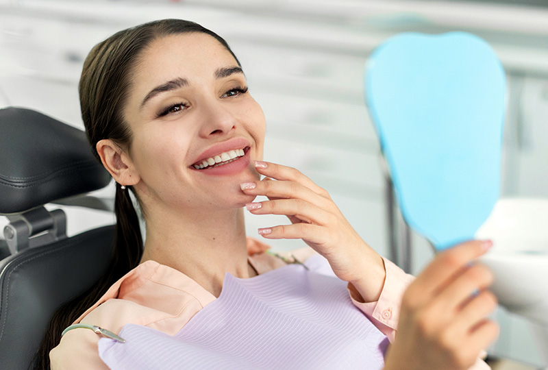 A woman is sitting in a dental chair, smiling and looking at her reflection in the mirror while holding up a blue toothbrush.