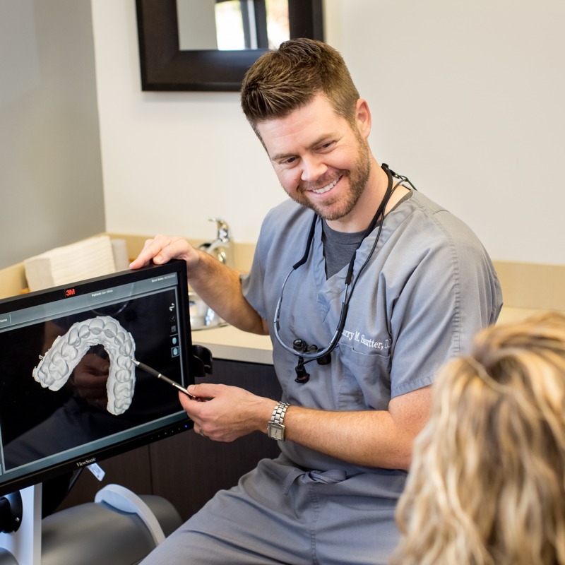 A dental professional, possibly a dentist or orthodontist, is demonstrating a 3D model of a human tooth to someone outside the frame.