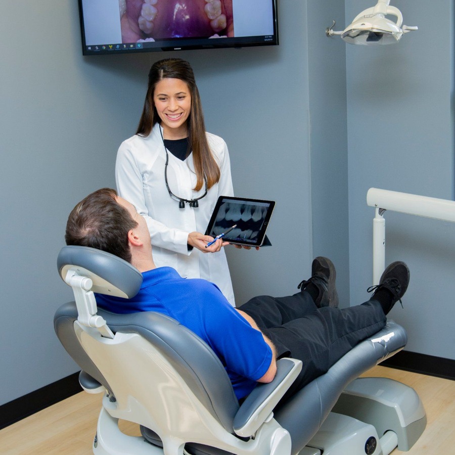 A dental professional in a white coat, standing beside a patient seated on a dental chair, demonstrating something on a tablet with an image of a mouth on the screen.