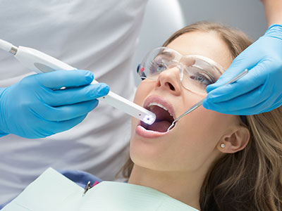 The image shows a dental professional performing a teeth cleaning procedure on a patient, with the patient wearing a blue surgical gown and having their mouth open for examination.